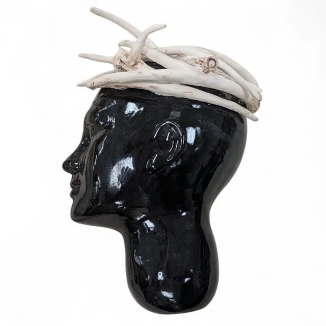 Side profile of a slip-cast head glazed glossy black, facing left. A crown of pale, bone-white antlers, tinged rust-brown with iron oxide, is tangled across the top of the head.
