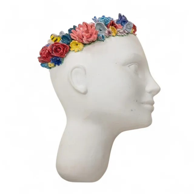 Side profile of an unglazed white porcelain head, facing right. A crown of glossy, brightly colored sculpted flowers in red, pink, blue, purple and yellow, with a small bumblebee among them, rings the top of the head.