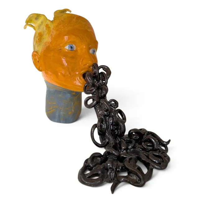 A glossy orange head with wide blue eyes, two swooping yellow tufts of hair, and a blue-gray neck. A tangled stream of dark brown, glossy clay coils and loops pours from its open mouth and spreads into a heap on the ground in front of it.