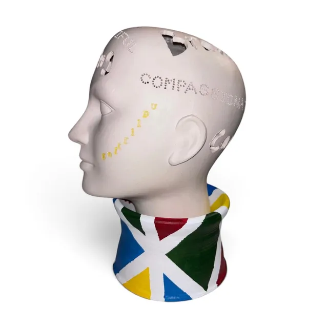 Side-profile ceramic head sculpture in matte white clay, facing left, with a smooth mannequin-like face. Words are cut into the skull as perforated dots and carved cutouts, including “COMPASSIONATE” across the temple and fragments of other words around the crown, with small yellow raised letters trailing diagonally across the cheek. The head sits in a flared collar base hand-painted with bold triangles in blue, red, green, and yellow, separated by white stripes.