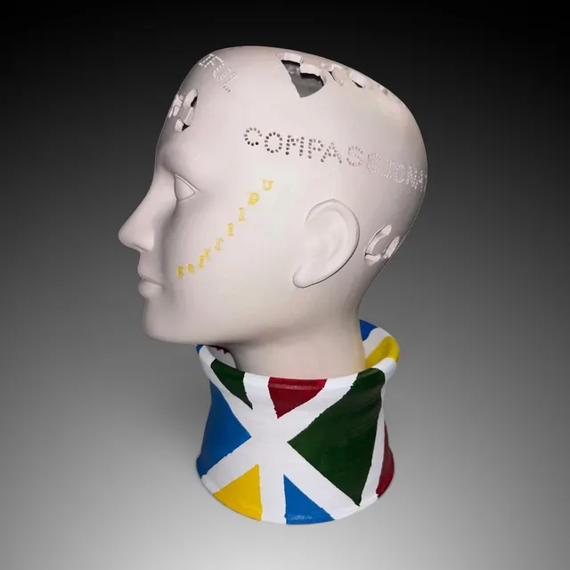 Side-profile ceramic head sculpture in matte white clay, facing left, with a smooth mannequin-like face. Words are cut into the skull as perforated dots and carved cutouts, including “COMPASSIONATE” across the temple and fragments of other words around the crown, with small yellow raised letters trailing diagonally across the cheek. The head sits in a flared collar base hand-painted with bold triangles in blue, red, green, and yellow, separated by white stripes.