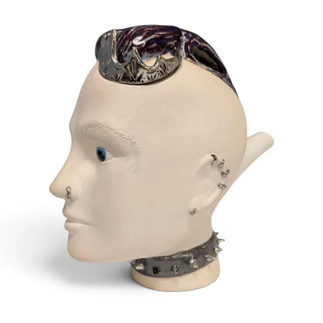 Side-profile ceramic teapot shaped like a head, facing left, in creamy off-white unglazed clay with a calm face and glossy blue eyes. The lid is a purple-and-white glazed mohawk topped with a metallic silver luster crown. A spout extends from the back of the head. Silver luster piercings line the ear and nose, and a gray spiked choker with silver luster studs circles the neck.