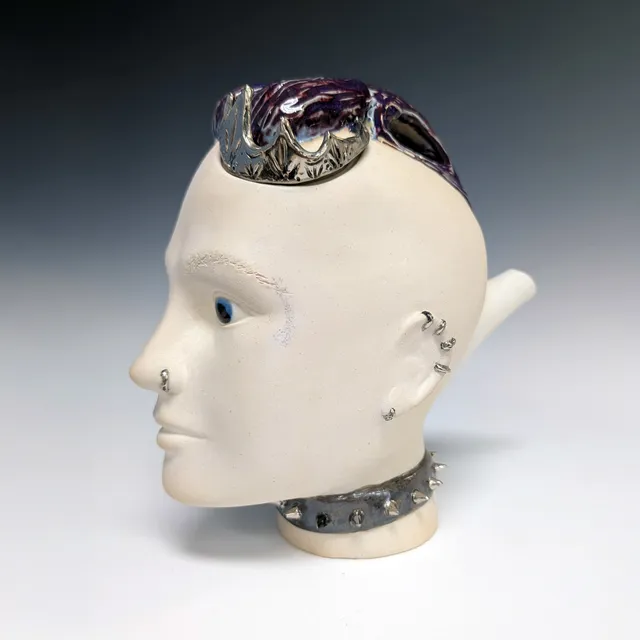 Side-profile ceramic teapot shaped like a head, facing left, in creamy off-white unglazed clay with a calm face and glossy blue eyes. The lid is a purple-and-white glazed mohawk topped with a metallic silver luster crown. A spout extends from the back of the head. Silver luster piercings line the ear and nose, and a gray spiked choker with silver luster studs circles the neck.