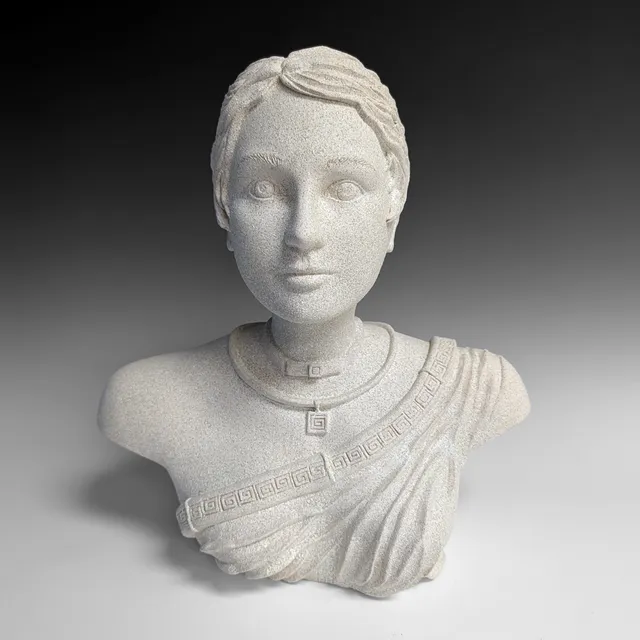 A bust of a woman with short swept-back hair and a calm, direct gaze, finished in a speckled stone texture. She wears a Greek key pendant on a choker and a draped garment across one shoulder, edged with a Greek key border.